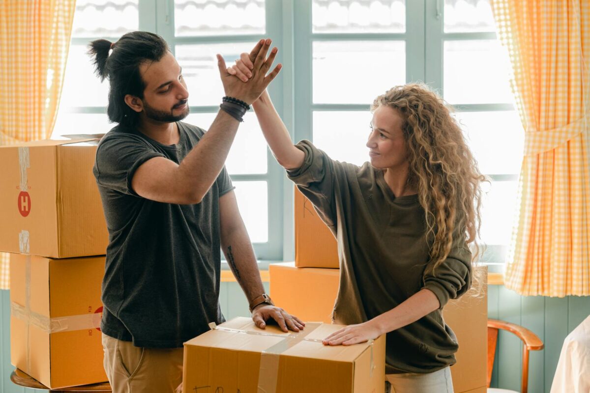 זוג חוגג מעבר לדירה חדשה עם קרטונים מסביב, מסמל הצלחה כלכלית כמו לקיחת הלוואה או משכנתא, ייעוץ פיננסי וחיסכון בכסף.