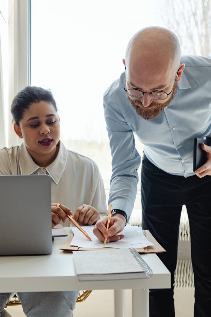 Two professionals working together at an office desk with a laptop and documents.